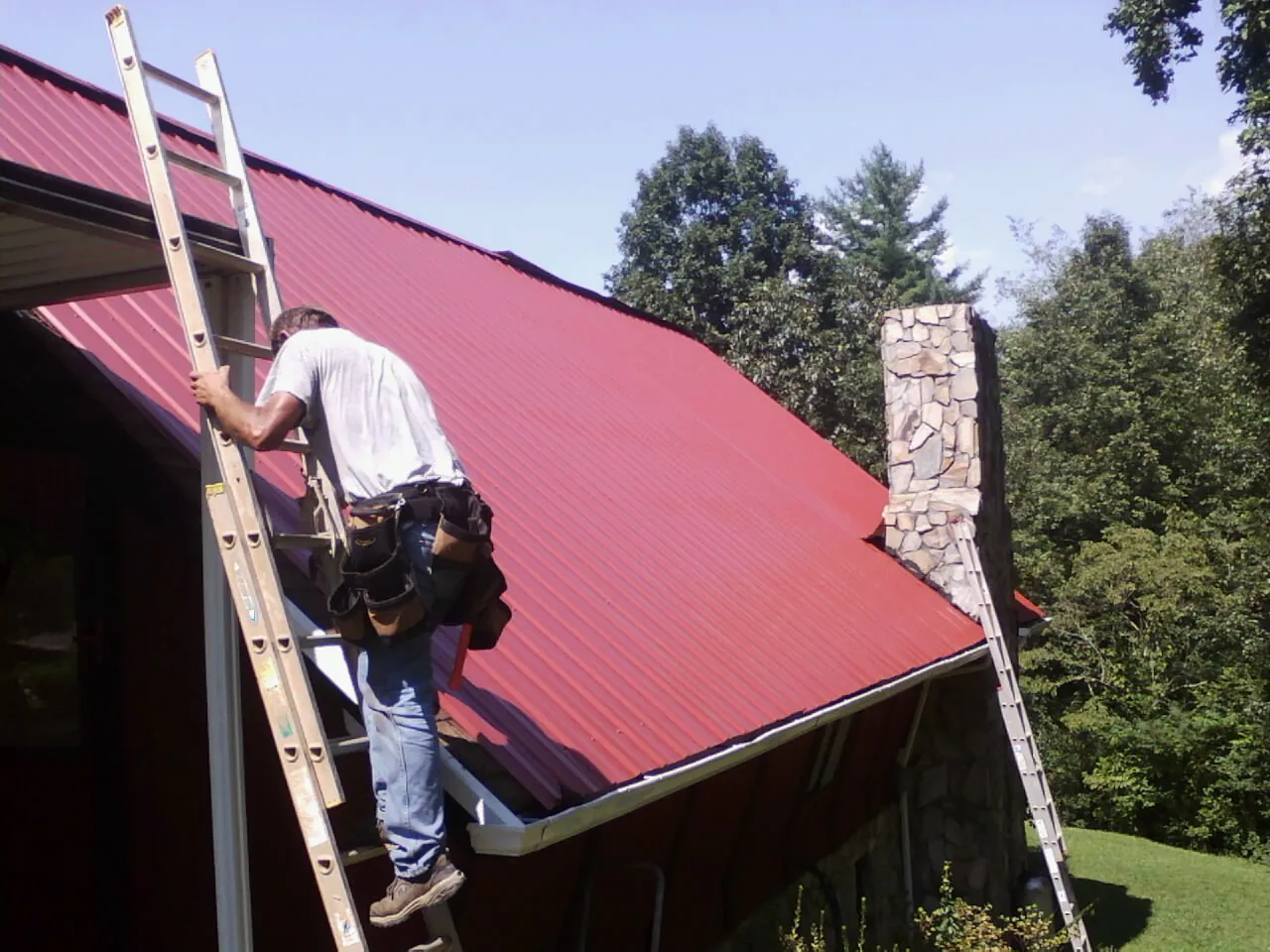 Roofer on a ladder beside a red metal roof and stone chimney — Soffit & Fascia Repair by Roofing Brothers in Gordonville